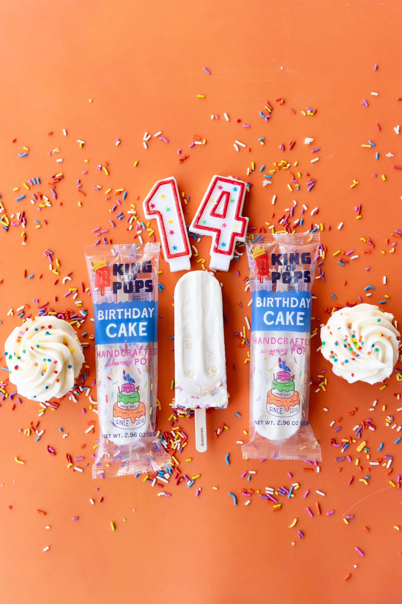 King of Pops Birthday Cake pops against an orange background, surrounded by cupcakes, sprinkles, and birthday cake candles.